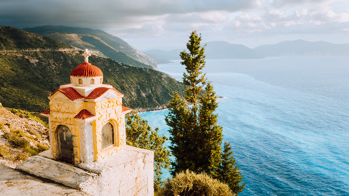Colorful Proskinitari shrine lantern on pedestal on the cliff edge. Amazing coastline view with cloudscape in the background